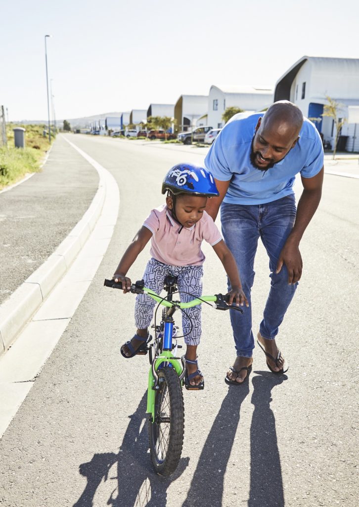 A father holds onto the seat of a bicycle, supporting his young son as he learns to ride, with Citra eHomes visible in the background.