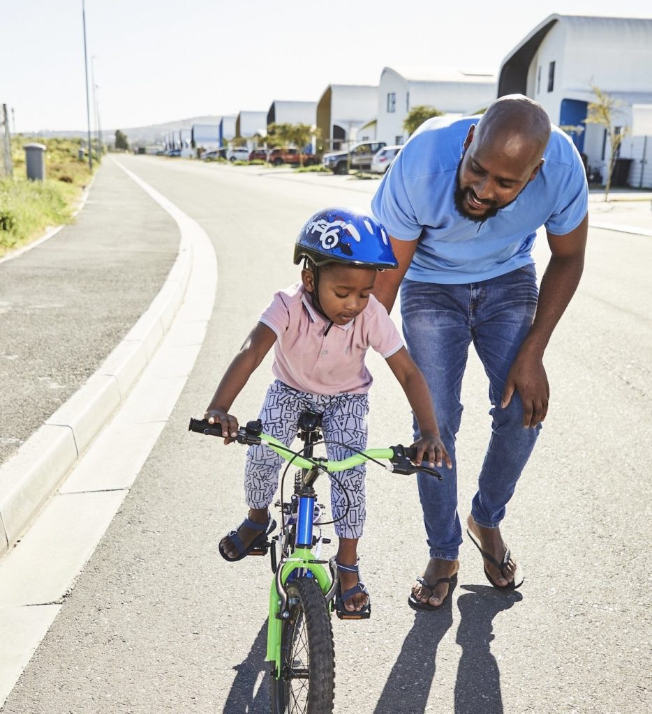 A father holds onto the seat of a bicycle, supporting his young son as he learns to ride, with Citra eHomes visible in the background.