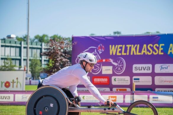 Side profile of Canadian Paralympic athlete Austin Smeenk at the starting line of a race. He is leaning forward in his specialized racing wheelchair, wearing a white helmet with the number 3. A large purple event banner is visible in the background.