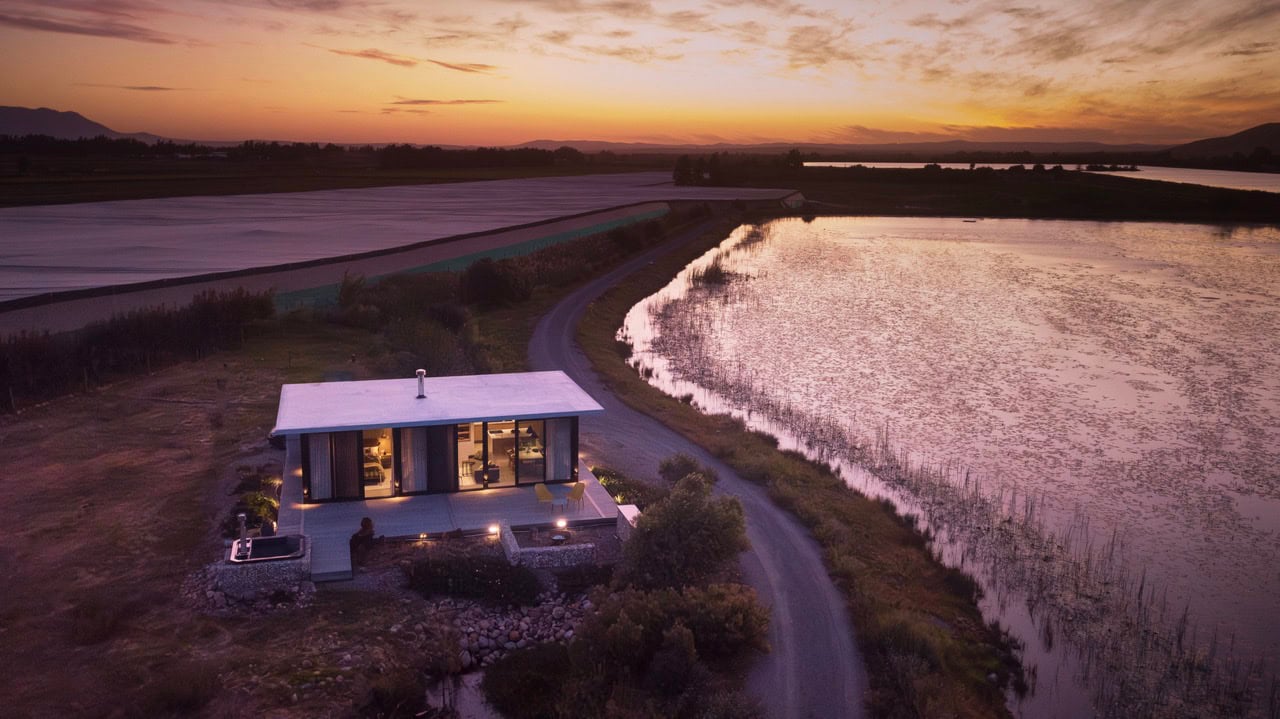 A lush, modern house situated near a serene water body at sunset, showcasing innovative architecture.