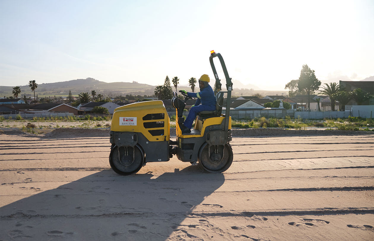 Worker operating a yellow road roller on a construction site in a sunny outdoor setting.